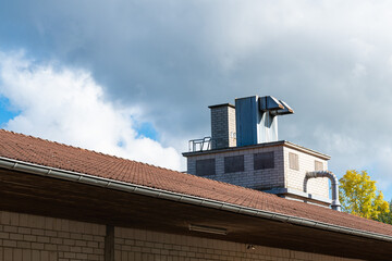 A chimney and metallic exhaust system are visible atop a building, set against a backdrop of clouds and blue sky, indicating industrial or commercial activity.