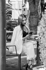 Obraz premium Stylish woman in a monochrome outfit seated at a cafe, engaged with her smartphone under natural light