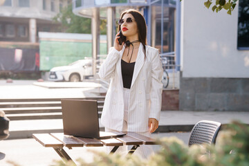Confident Businesswoman in Striped Suit Making a Phone Call Outdoors with Laptop on Table
