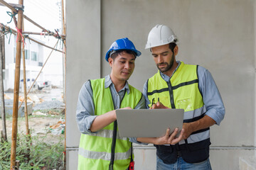 Young engineer with laptop computer explain to foreman about a floor plan. Both wear construction helmet and safety vest. Work environment at the construction site of housing projects.