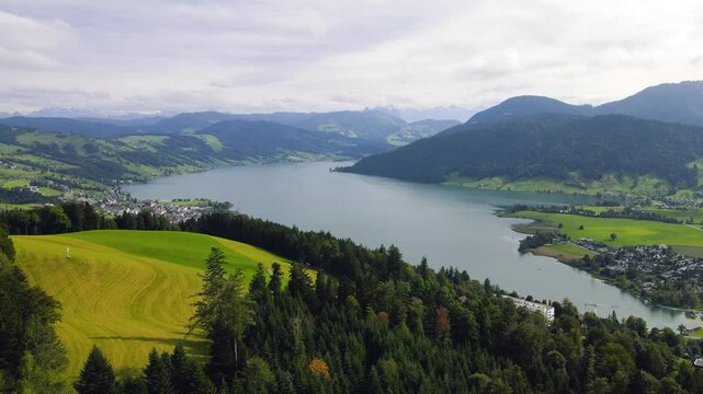 A cinematic aerial shot of a stunning glacial lake surrounded by alpine villages on its shores with the dramatic Swiss rocky mountains in the background featuring Lake Aegeri in switzerland 






