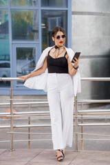 Fototapeta premium Stylish Young Woman in Chic Outfit with Sunglasses and Mobile Phone Outdoors
