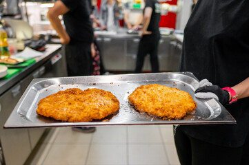 A chef’s hands in black gloves holding a metal tray with two large, golden-brown schnitzels in a professional kitchen. Blurred chefs in the background. Perfect for culinary, food preparation