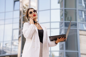 Professional Businesswoman in Stylish Striped Suit Using Laptop and Phone Outside Modern Office...