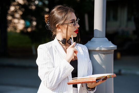 A Young Businesswoman in Striped Blazer Thinking Pensively While Holding a Notebook and Pen Outdoors