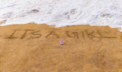 It's a boy and it's a girl baby written text announcement on the beach sand with shoe. Clean sea water and small beach waves are in background