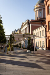 Naklejka premium Street and old buildings in the center of Moscow. In the middle of the street there are wooden supports, entwined with flowers. There is a scooter, benches.