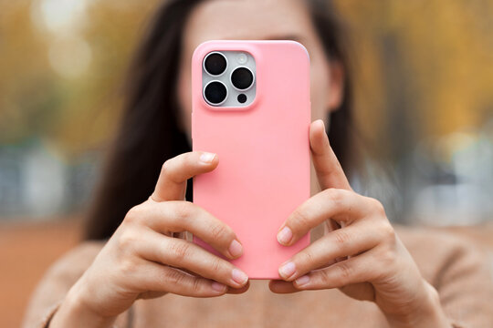 Closeup mobile phone in pink silicone case in girl hands, no face. Woman in fall city park make a photo on modern new smartphone.