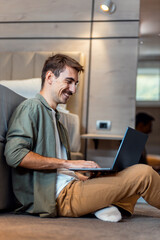 Man is sitting next to the bed in bedroom on the floor using a laptop.