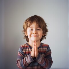 A joyful child with clasped hands smiles warmly, set against a simple backdrop that highlights his pure innocence and happiness.