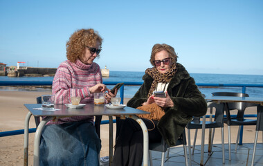 Mother and daughter using smartphones while enjoying coffee by the beach