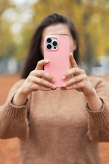 Vertical shot young woman holding smartphone in pink silicone case outdoors. Teen girl take a picture by mobile phone in autumn city park. 