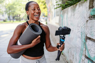 Sporty female vlogger holding her camera and exercise mat in the city