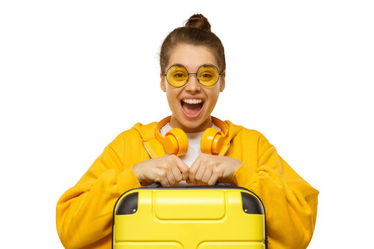 Happy excited girl wearing hoodie, yellow colored glasses and headphones around neck, holding travel suitcase, feeling ready for flight