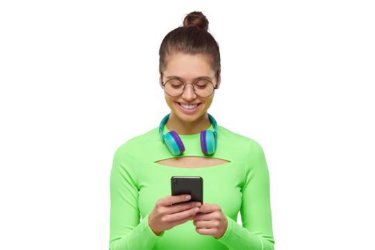Young girl in glasses and neon top, with headphones around neck, holding phoen in hands, reading and texting messages with happy smile