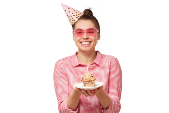 Excited funny young woman in pink glasses and birthday hat, holding cake on plate with two hands, making wish with closed eyes