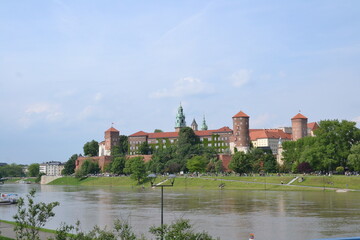 A scenic view of Wawel Castle in Kraków, Poland, showcasing its historic red-brick architecture, green domes, and surrounding park along the Vistula River under a clear sky.