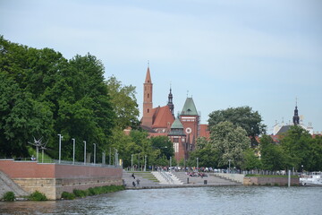 The photo shows a serene riverside scene in Wrocław, with lush green trees along the promenade. In...