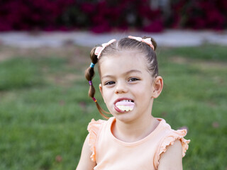 A young girl with pigtails smiles while playfully chewing a sweet. She sits on green grass at the park.