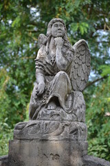 The image depicts a stone statue of an angel sitting thoughtfully, resting her chin on her hand. The angel has detailed wings, flowing garments, and a serene expression, surrounded by greenery.