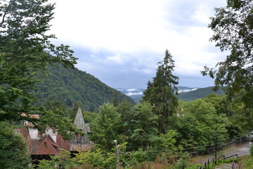 This photo captures a serene mountain landscape with dense green forests and mist-covered hills in the background. In the foreground, old buildings with pointed roofs are nestled among the trees.