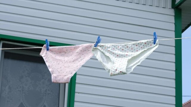 Two pairs of floral-patterned panties hanging on a clothesline in front of a house window, drying in the fresh air. The scene reflects simplicity, care, and the natural process of air drying clothes.