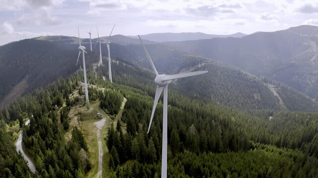 Wind turbines on a forested mountain, set against a bright sky with clouds, representing renewable energy in nature.
