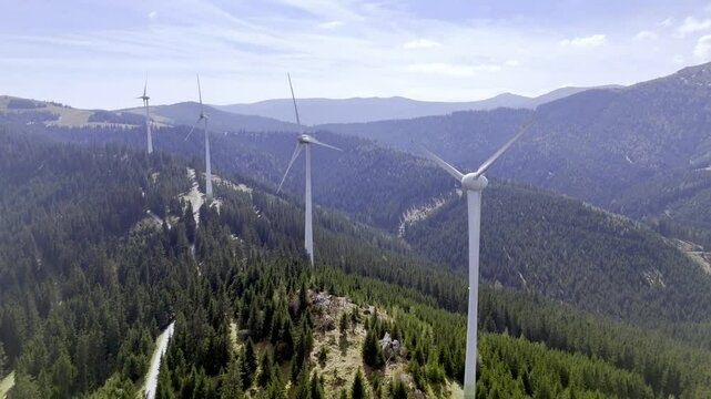Wind turbines on a forested mountain, set against a bright sky with clouds, representing renewable energy in nature.
