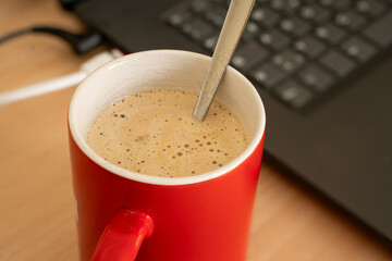 Red cup of coffee with crema and spoon in the cup standing besides a notebook. Just keyboard and some plugs can be seen from the notebook. Close up.