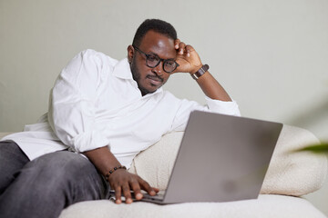 Handsome Black African American Man Working on Laptop Computer while Sitting on a Sofa in Cozy Living Room. Freelancer Working From Home. Browsing Internet, Using Social Networks, Having Fun in Flat.