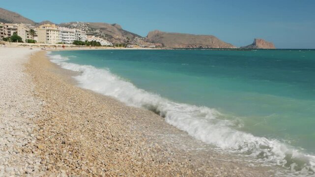 Turquoise Waves Lapping Against Pebble Shore Create Tranquil Scene, While Distant Buildings and Mountains Enhance Beauty of Mediterranean Landscape