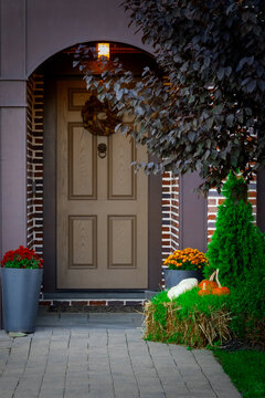 A cozy front entrance features a wooden door adorned with a wreath, flanked by brick walls and decorative plants. Pumpkins and a hay bale add an autumnal touch to the neatly arranged garden area.