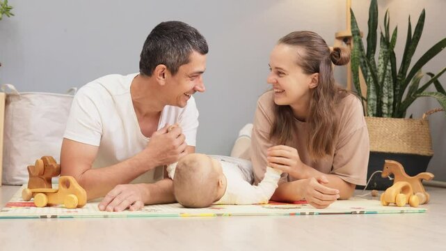 Smiling cheerful mother and father lying on floor with infant interacting with toddler child together enjoying babyhood moments of care and development in living room at home