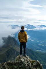 A lone figure in a hoodie stands on a cliff, taking in the vast expanse of mountains under a cloudy sky.