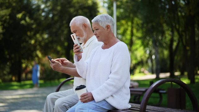 Charming senior couple with white hair happily relaxing in city park, sitting on bench, oblivious to each other, enjoying smartphones on beautiful sunny day. Concept of happy retirement life.