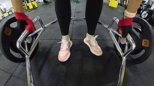 A Woman prepares for a Deadlift exercise by securing wrist straps to the Barbell loaded with 20 kg plates. The close-up, first-person perspective emphasizes the focus on form and grip strength.