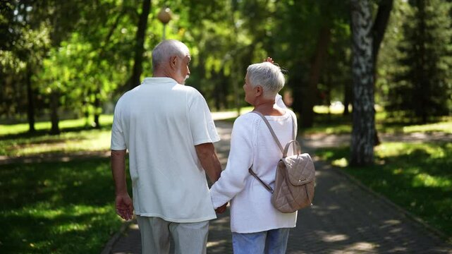 Back view of unrecognizable senior couple leisurely walking through sunny park, holding hands and savoring each others company under the lush trees, having active retirement lifestyle, slow motion.
