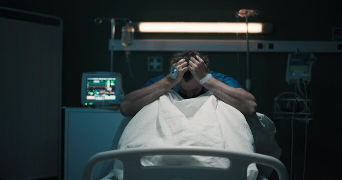 Exhausted man in hospital ward suffering from migraine shaking holding head with hands.