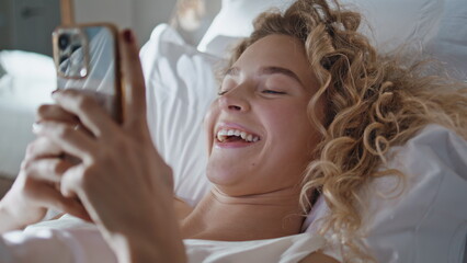Naklejka premium Joyful woman lying smartphone in cozy bed closeup. Happy curly girl browsing web