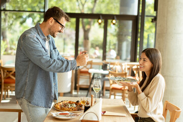 Couple enjoying a cozy meal together in a bright cafe, capturing memories with a smartphone during a sunny afternoon