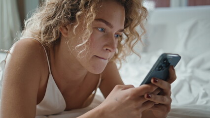 Cheerful girl texting smartphone in home bed. Closeup curly woman enjoying web