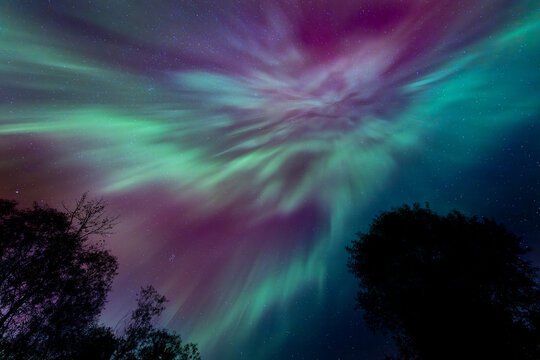 Northern Lights erupt over a Minnesota lake in the dark sky overhead shining a bright rainbow of Aurora light and colors over the forests and clean water during strong solar storm