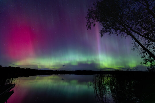 Northern Lights erupt over a Minnesota lake in the dark sky overhead shining a bright rainbow of Aurora light and colors over the forests and clean water during strong solar storm