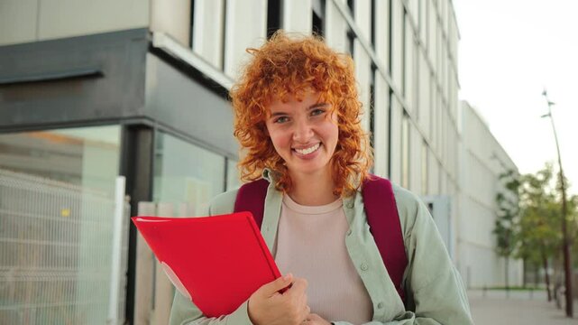 Smiling redhead student looking at the camera while holding a red folder and wearing a backpack, standing outdoors in an urban setting, showing confidence and a positive attitude on a bright day.