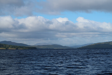 A tranquil Scottish landscape showcasing a loch with gentle ripples. Rolling hills and mountains stand tall in the background, topped by a partly cloudy sky with hints of blue peeking through.