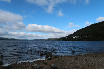 A serene Scottish lakeside scene with a rocky shore, calm blue waters, and a distant hilly landscape. The sky is partly cloudy with patches of blue, and white buildings are visible on the hill.