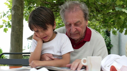 Elderly man and young boy reading a book together outdoors, with the boy showing a puzzled expression as they share a moment of learning and bonding under the shade of a tree