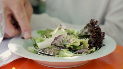 Close-up of a fresh and colorful mixed salad with lettuce leaves on a white plate, held by a person with a fork, ready to eat at an outdoor cafe