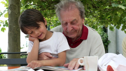 Elderly man and young boy reading a book together outdoors, with the boy showing a puzzled expression as they share a moment of learning and bonding under the shade of a tree