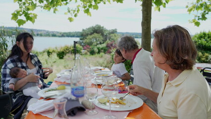 Family enjoying a meal outdoors with lake views, featuring a mother holding her baby, a young boy, and grandparents, all savoring the moment together